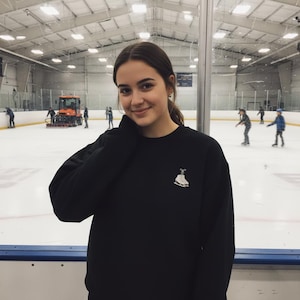 以下が含まれることがあります： 白いスケート靴のグラフィックが入った黒いスウェットシャツ。スケートリンクで、このスウェットシャツを着た人がいます。背景には他の人がスケートをしています。