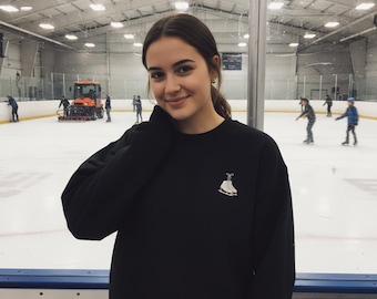 May include: A black sweatshirt with a white ice skate graphic. The sweatshirt is worn by a person in an ice skating rink. Other people are ice skating in the background.