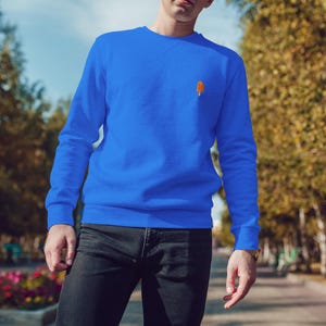 Könnte beinhalten: Ein königsblaues Sweatshirt mit einer kleinen orangefarbenen Eis am Stiel-Grafik auf der Brust. Dazu wird eine schwarze Jeans getragen. Der Hintergrund ist verschwommen mit Bäumen und einem Weg.