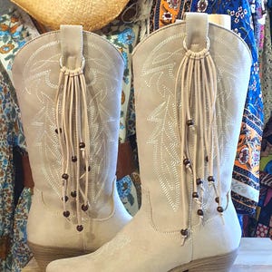 Puede incluir: Un par de botas beige a media pantorrilla con costuras decorativas blancas, punta puntiaguda y tacón de bloque. Cada bota tiene un anillo de metal circular en la parte superior, del que cuelgan flecos de ante beige con pequeñas cuentas marrones.