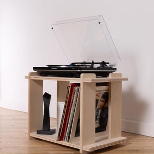K&ouml;nnte beinhalten: Ein helles Sperrholz-Plattenspieler-Regal mit einer rechteckigen oberen und unteren Ablage. Das Regal tr&auml;gt einen schwarzen Audio-Technica-Plattenspieler mit transparenter Abdeckung sowie mehrere Schallplatten und eine schwarze Skulptur.