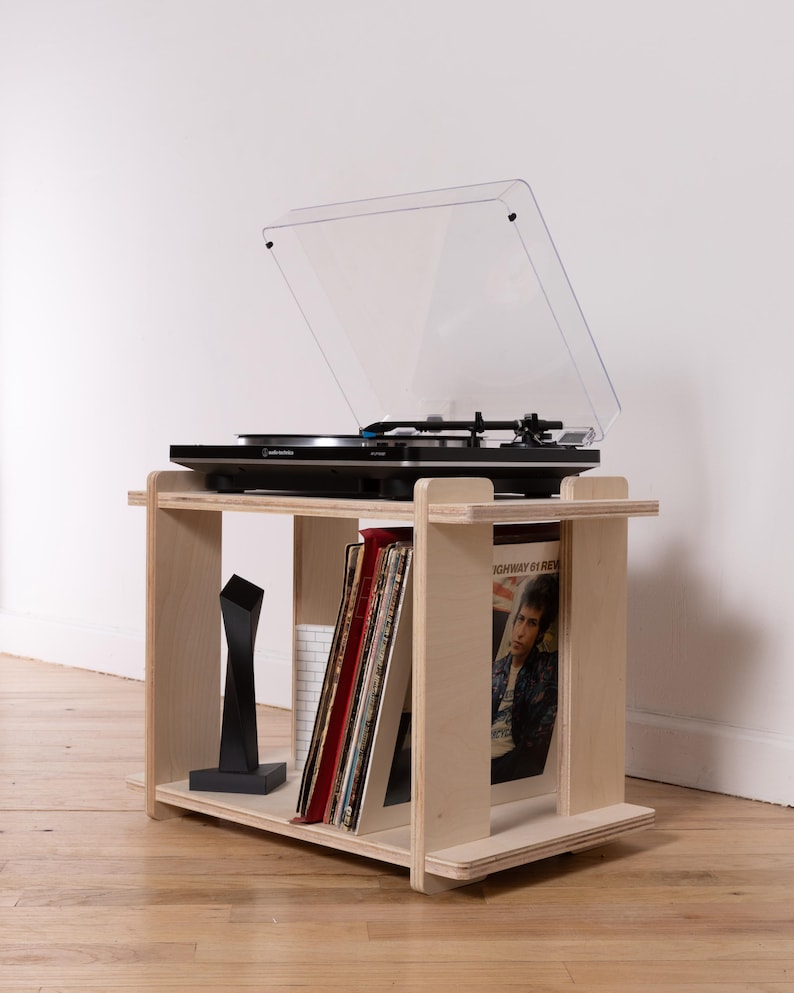 K&ouml;nnte beinhalten: Ein helles Sperrholz-Plattenspieler-Regal mit einer rechteckigen oberen und unteren Ablage. Das Regal tr&auml;gt einen schwarzen Audio-Technica-Plattenspieler mit transparenter Abdeckung sowie mehrere Schallplatten und eine schwarze Skulptur.