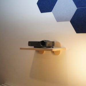 May include: A black projector sits on a small, light-colored wooden shelf attached to a white wall. The shelf has rounded supports. Above, there are blue and gray hexagonal acoustic panels.