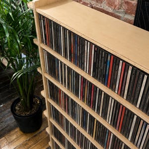 May include: A light-colored wooden CD storage shelf filled with compact discs. The shelf has multiple horizontal levels and is positioned next to a potted plant. The background features a brick wall.