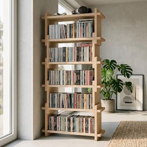 May include: A tall, light-colored wooden shelving unit with five horizontal tiers, constructed with interlocking side panels. The shelves are filled with numerous compact discs arranged vertically, and a pair of headphones rests on the top shelf.