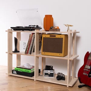 K&ouml;nnte beinhalten: Ein helles Sperrholzregal mit drei Ebenen, das einen schwarzen Plattenspieler, eine orangefarbene Vase, einen gelben Gitarrenverst&auml;rker, Schallplatten, B&uuml;cher mit den Titeln Virgil Abloh ICONS und Tom Ford, zwei Kameras und eine kleine metallische Vogelfigur enth&auml;lt.