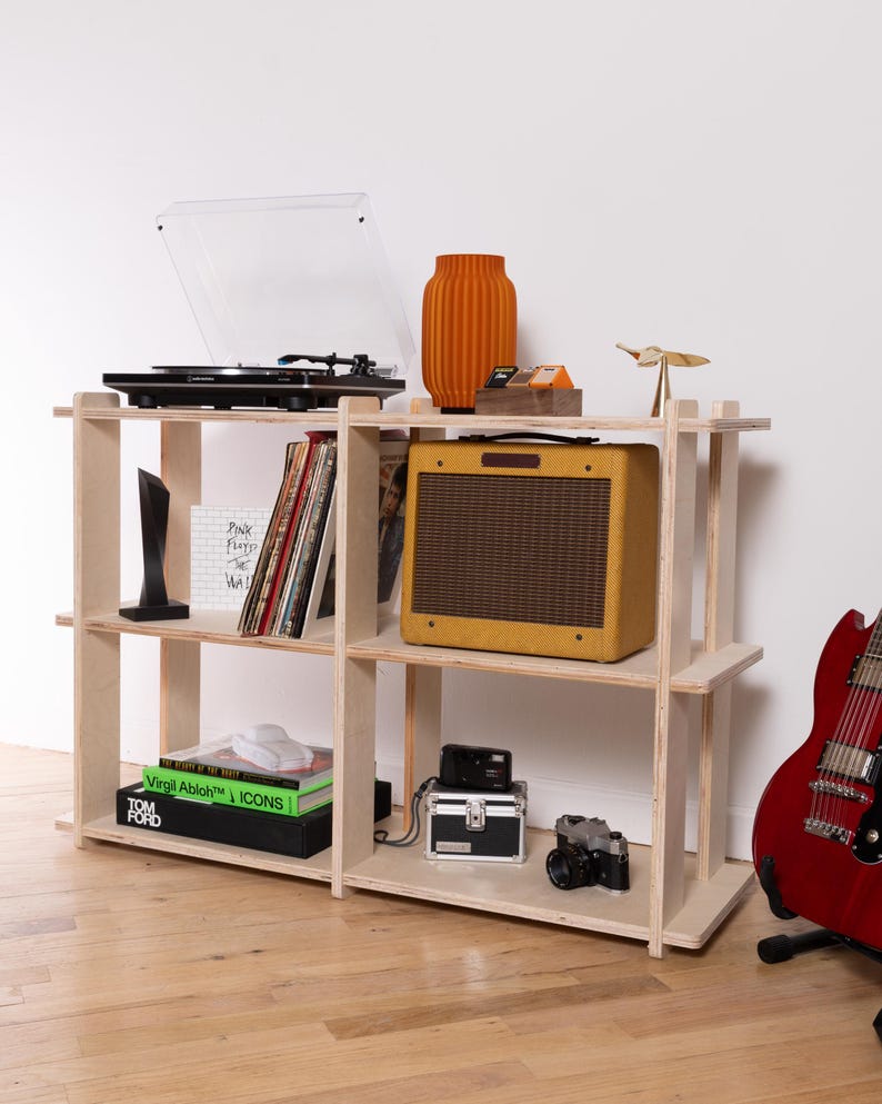 K&ouml;nnte beinhalten: Ein helles Sperrholzregal mit drei Ebenen, das einen schwarzen Plattenspieler, eine orangefarbene Vase, einen gelben Gitarrenverst&auml;rker, Schallplatten, B&uuml;cher mit den Titeln Virgil Abloh ICONS und Tom Ford, zwei Kameras und eine kleine metallische Vogelfigur enth&auml;lt.
