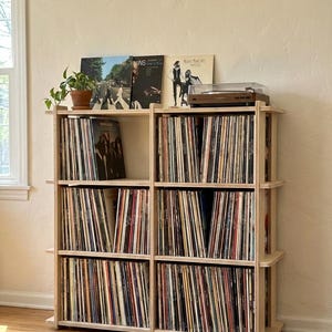 K&ouml;nnte beinhalten: Ein helles Holzregal zur Aufbewahrung von Schallplatten mit drei Ebenen, unterteilt in sechs F&auml;cher, gef&uuml;llt mit Vinyl-Schallplatten. Ein Plattenspieler steht oben rechts und eine Topfpflanze steht oben links. Mehrere Schallplattencover sind aufrecht ausgestellt.