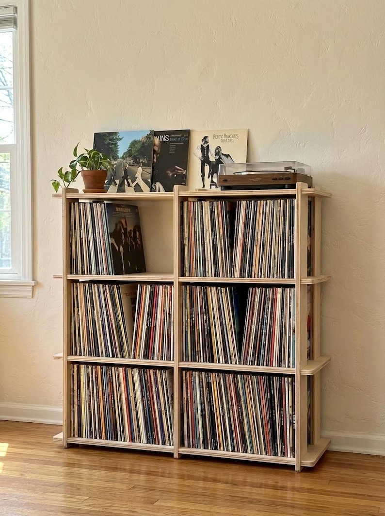 K&ouml;nnte beinhalten: Ein helles Holzregal zur Aufbewahrung von Schallplatten mit drei Ebenen, unterteilt in sechs F&auml;cher, gef&uuml;llt mit Vinyl-Schallplatten. Ein Plattenspieler steht oben rechts und eine Topfpflanze steht oben links. Mehrere Schallplattencover sind aufrecht ausgestellt.