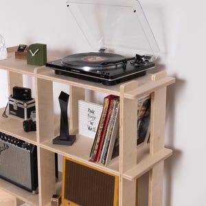 May include: A black turntable with a clear cover sits on a light wood shelf unit. The shelf displays vinyl records, a small amplifier, a clock, and other decorative items. The text "Pink Floyd The Wall" is visible.