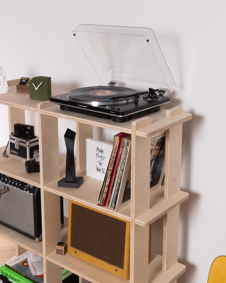 K&ouml;nnte beinhalten: Ein helles Holzregal mit einem schwarzen Audio-Technica-Plattenspieler mit durchsichtiger Abdeckung, einer schwarzen abstrakten Skulptur, einem Pink Floyd The Wall-Album, einem gelben Gitarrenverst&auml;rker sowie weiteren Elektronikartikeln und B&uuml;chern.