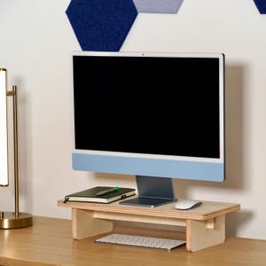 May include: A computer monitor with a light blue base sits on a wooden monitor stand. A keyboard, mouse, notebook, and pen are on the stand. The monitor is on a wooden desk. A gold lamp is on the left.
