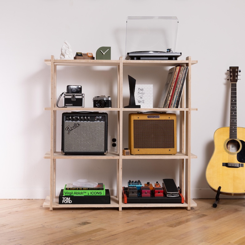 K&ouml;nnte beinhalten: Ein helles Holzregal mit vier Ebenen, das einen Plattenspieler, Kameras, einen schwarzen Fender-Verst&auml;rker, einen gelben Verst&auml;rker, B&uuml;cher und Gitarrenpedale enth&auml;lt. Eine helle Akustikgitarre steht rechts auf einem Bodenst&auml;nder.