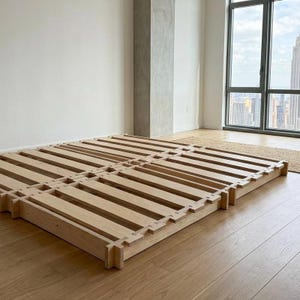 以下が含まれることがあります： スラットデザインの明るい色の木製ベッドフレーム。フレームは複数のセクションで構成されており、床に直接置かれています。部屋には、街の景色を望む大きな窓と、広葉樹のフローリングがあります。