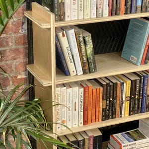 May include: A wooden bookshelf with numerous books of various sizes and colours. Titles such as "Sapiens", "The Alchemist", and "Pride and Prejudice" are visible. The bookshelf has a simple design with multiple shelves, ideal for home or office use.