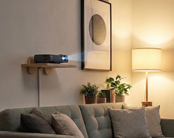 May include: A light-coloured wooden wall-mounted shelf holding a black digital projector. The projector is positioned to cast a light beam towards the right side of the room, above a grey tufted sofa with several throw pillows and potted plants.