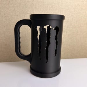 Puede incluir: Taza negra con asa y diseño de garras. La taza cilíndrica tiene una base ancha y un asa robusta. Las marcas de garras están recortadas de la taza, revelando el interior.