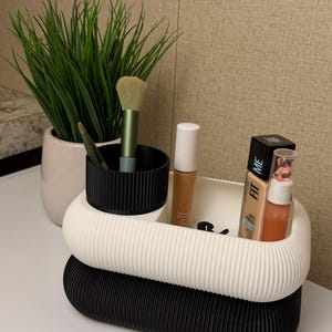 Puede incluir: Un organizador de maquillaje de tres niveles en negro, blanco y negro, con un pequeño soporte cilíndrico negro para brochas. Contiene brochas de maquillaje, base y otros productos de belleza. Una pequeña planta en maceta está en el fondo.