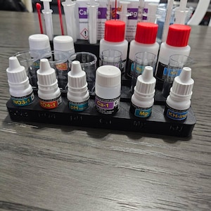 Op de afbeelding: Een zwarte plastic organizer met geëtiketteerde flessen met vloeibare aquariumtestreagentia. De flessen zijn geëtiketteerd met: NO3-1, PO4-1, PH, Ca-1, Mg, KH-ing. De organizer bevat ook rode en doorzichtige druppelaars.