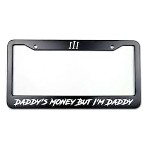 May include: Black license plate frame with white text that reads "DADDY'S MONEY BUT I'M DADDY" in a stylized font. The top of the frame has the Roman numeral "III".