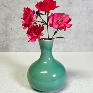 May include: A small, round, teal-colored ceramic vase holding a small bouquet of vibrant pink roses. The vase has a glossy finish and a narrow neck. The roses are in full bloom, with multiple petals.
