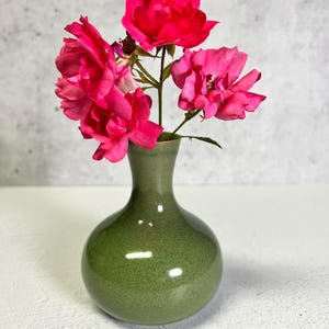 May include: A small, green ceramic vase holding a few vibrant pink roses. The vase has a rounded body and a narrow neck. The roses are in full bloom, with multiple petals and a rich, deep pink color. The background is a neutral, light gray.