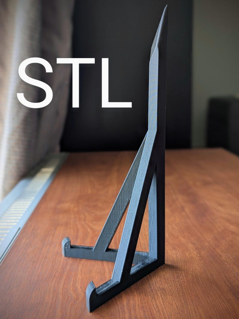 May include: A black, geometric stand with the letters "STL" in white. The stand is made of dark gray material and has a triangular design. It is placed on a wooden surface.