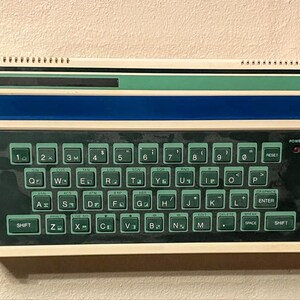 May include: A vintage white and blue computer with a green keyboard. The keyboard has keys labelled with letters, numbers, and symbols. The computer has a small screen at the top.