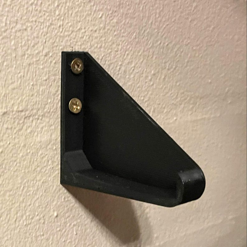 May include: A black plastic wall mount with two gold screws. The mount is angled and has a small shelf.
