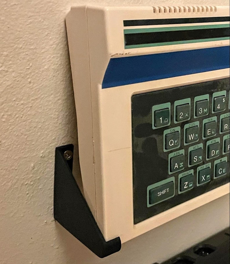 May include: A white calculator with a black and blue display and a black keyboard with keys labelled with mathematical functions, such as SIN, COS, TAN, LOG, and more. The calculator is mounted on a black wall mount.