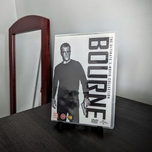 May include: A DVD case for "Bourne: The Ultimate 5-Movie Collection" featuring a black and white image of Matt Damon. The case is displayed on a black stand, with the title and movie details visible. The background includes a dark wooden table and a mirror.