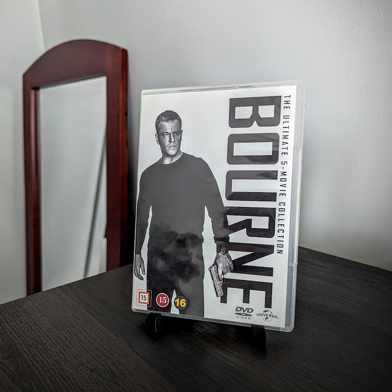 May include: A DVD case for "Bourne: The Ultimate 5-Movie Collection" featuring a black and white image of Matt Damon. The case is displayed on a black stand, with the title and movie details visible. The background includes a dark wooden table and a mirror.