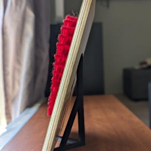 May include: A wooden plaque with a red, textured element, supported by a black metal stand. The plaque is made of light-colored wood and is angled, showcasing the red texture. The stand is a simple, geometric design.