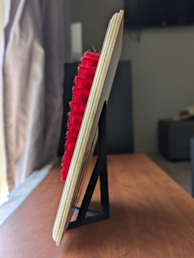 May include: A wooden plaque with a red, textured element, supported by a black metal stand. The plaque is made of light-colored wood and is angled, showcasing the red texture. The stand is a simple, geometric design.