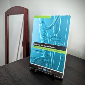 May include: A textbook titled "Making the Connections&sup3;" on a black stand. The cover is blue with a diagram of lab equipment. The text reads "A How-To Guide for Organic Chemistry Lab Techniques" by Anne B. Padias. The book is on a dark wooden surface.