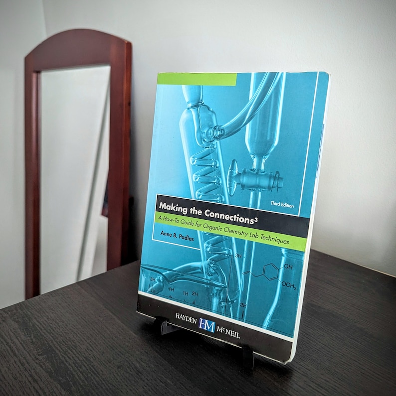 May include: A textbook titled "Making the Connections&sup3;" on a black stand. The cover is blue with a diagram of lab equipment. The text reads "A How-To Guide for Organic Chemistry Lab Techniques" by Anne B. Padias. The book is on a dark wooden surface.