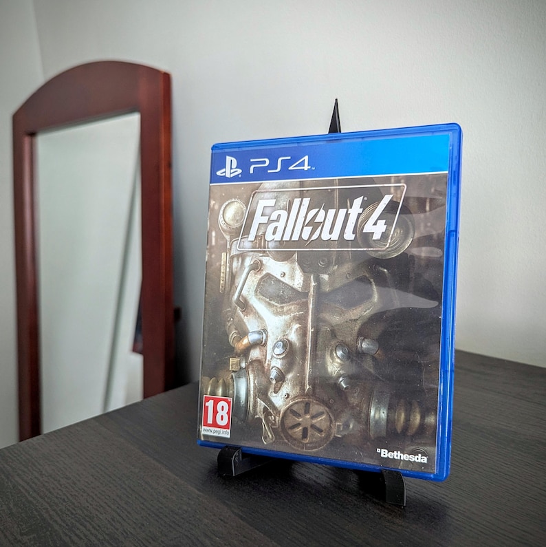 May include: A blue PS4 game case for Fallout 4, displayed on a black stand. The game cover features a detailed illustration of a futuristic helmet and the game title. The case is rated 18 and includes the Bethesda logo.