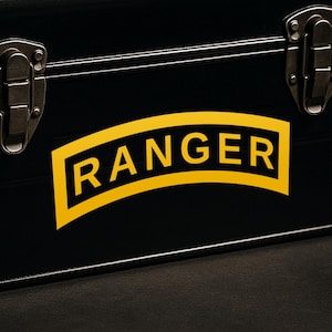 May include: Black metal storage box with a yellow and black "RANGER" emblem. The emblem is a yellow rectangle with the word "RANGER" in black, arched across the top. The box has metal latches and hinges.