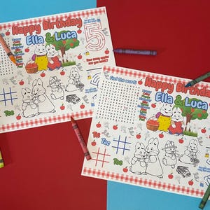 May include: Two birthday placemats with the text "Happy Birthday Ella & Luca" and illustrations of cartoon characters, apples, and a maze. Crayons in various colors are scattered around the placemats.