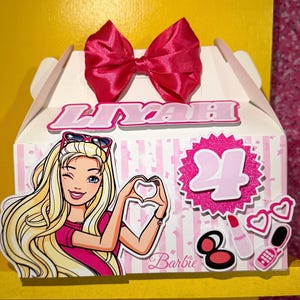 May include: A white and pink striped box with a large pink bow on top. The box features a Barbie graphic, the name "LIYAH", and the number "24". Additional decorative elements include makeup and heart-shaped glasses.