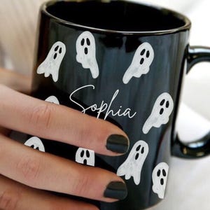 May include: Black ceramic mug decorated with white ghost illustrations. The mug has a black handle and the name "Sophia" in white script. The mug is held by a person with black nail polish.
