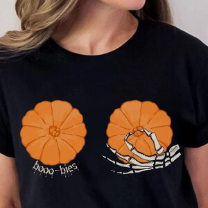 May include: Black t-shirt featuring two orange pumpkin illustrations. One pumpkin has the word "booo-bies" below it, and the other has a skeleton hand reaching for it. The shirt is a Halloween-themed design.