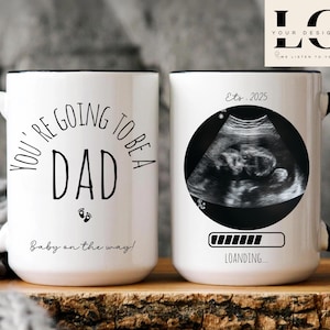 Puede incluir: Dos tazas de cerámica blanca con asas negras. Una taza tiene el texto "You're going to be a Dad" con un gráfico de huella de bebé y el texto "Baby on the way!" La otra taza tiene una imagen de ultrasonido en blanco y negro con el texto "Ets. 2025" y una barra de progreso con el texto "Loading..."