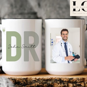 Puede incluir: Dos tazas de cerámica blanca con asas negras. Una taza presenta el texto "DR John Smith" con un "DR" verde y gris. La otra taza tiene una foto de un hombre con una bata blanca de médico.