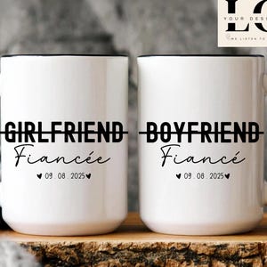 Puede incluir: Dos tazas de cerámica blanca con asas negras y letras negras. La taza de la izquierda dice "Girlfriend - Fiancée 09.08.2025" y la taza de la derecha dice "Boyfriend - Fiancé 09.08.2025".