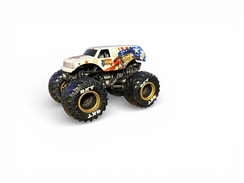 May include: A white monster truck toy with a patriotic theme. The truck features a design with red, white, and blue accents, stars, and a cartoon character. The large black tires have gold rims and the text "BKT".