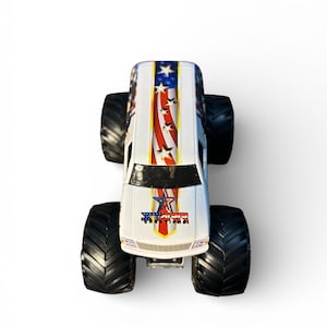 May include: A white toy monster truck with black tires. The truck features a patriotic design with red and white stripes, blue stars, and the word "STAR" in a star-shaped emblem on the front.
