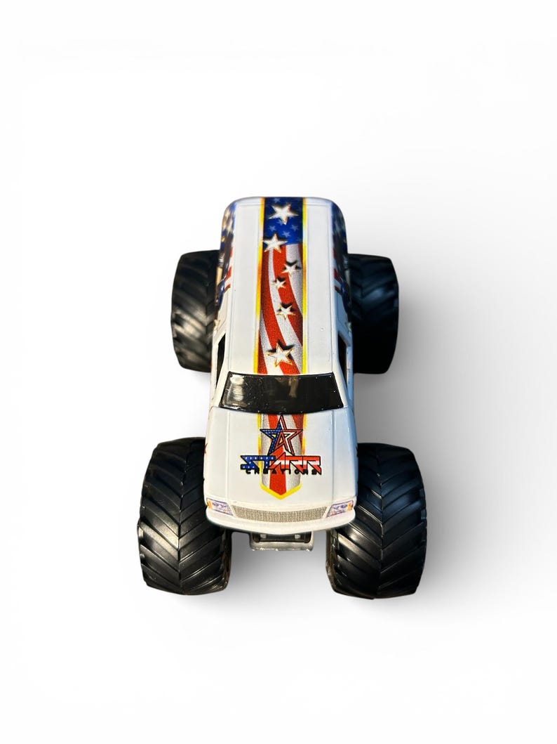 May include: A white toy monster truck with black tires. The truck features a patriotic design with red and white stripes, blue stars, and the word "STAR" in a star-shaped emblem on the front.