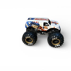 May include: A white monster truck toy with patriotic graphics, including stars and stripes, and a cartoon character. The truck has large black tires with gold rims and the text "BKT".