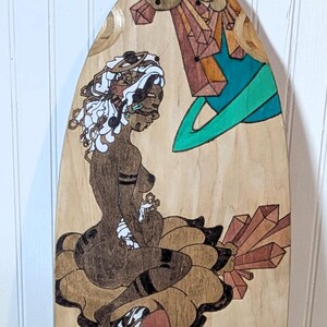 May include: A wooden skateboard deck with a hand-painted design featuring a woman with white hair sitting on a cloud, surrounded by geometric shapes and abstract patterns in brown, black, white, and green.